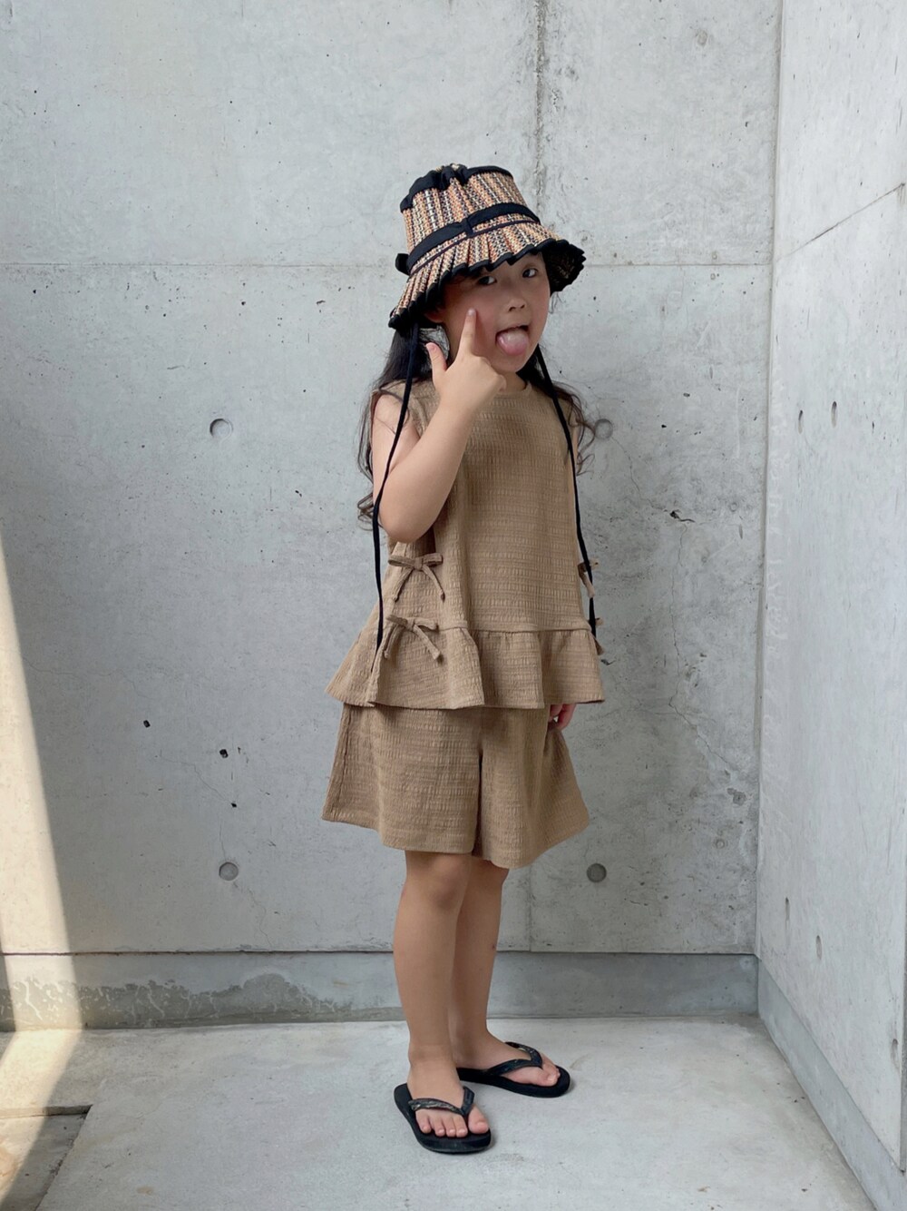 ブラウン系のパンツドレス、ブラック系のサンダル、ブラック系の帽子を着用したキッズの夏コーディネートの1枚目の写真