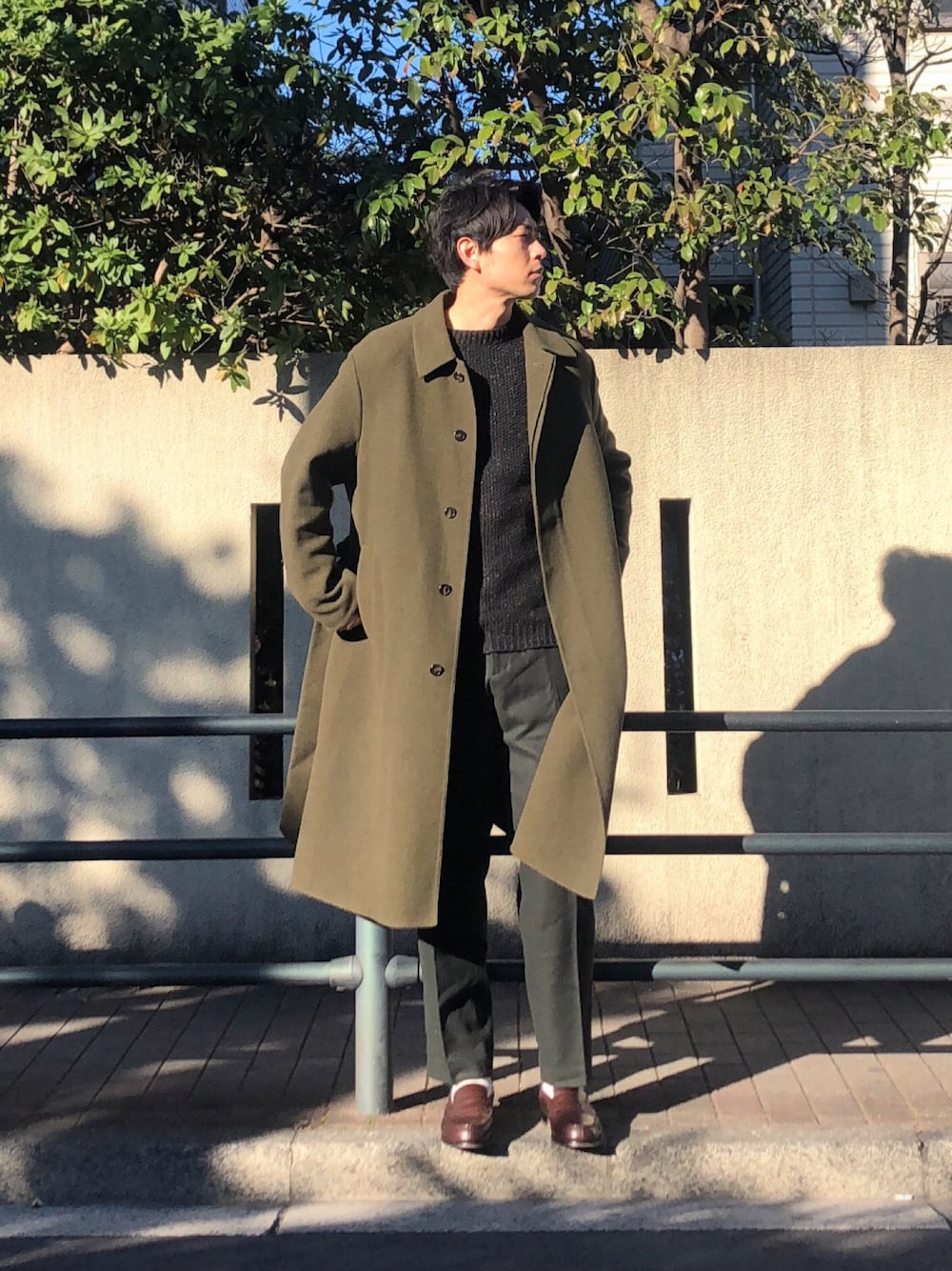 グリーン系のステンカラーコート、ブラック系のニット/セーター、グリーン系のスラックスを着用したメンズの冬コーディネートの1枚目の写真