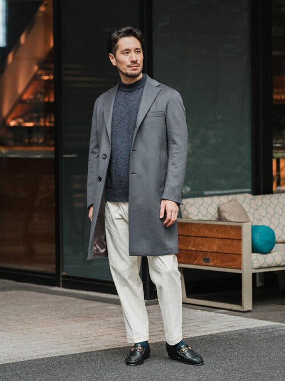 グレー系のチェスターコート、ブルー系のニット/セーター、ホワイト系のスラックスを着用したメンズの冬コーディネートの1枚目の写真