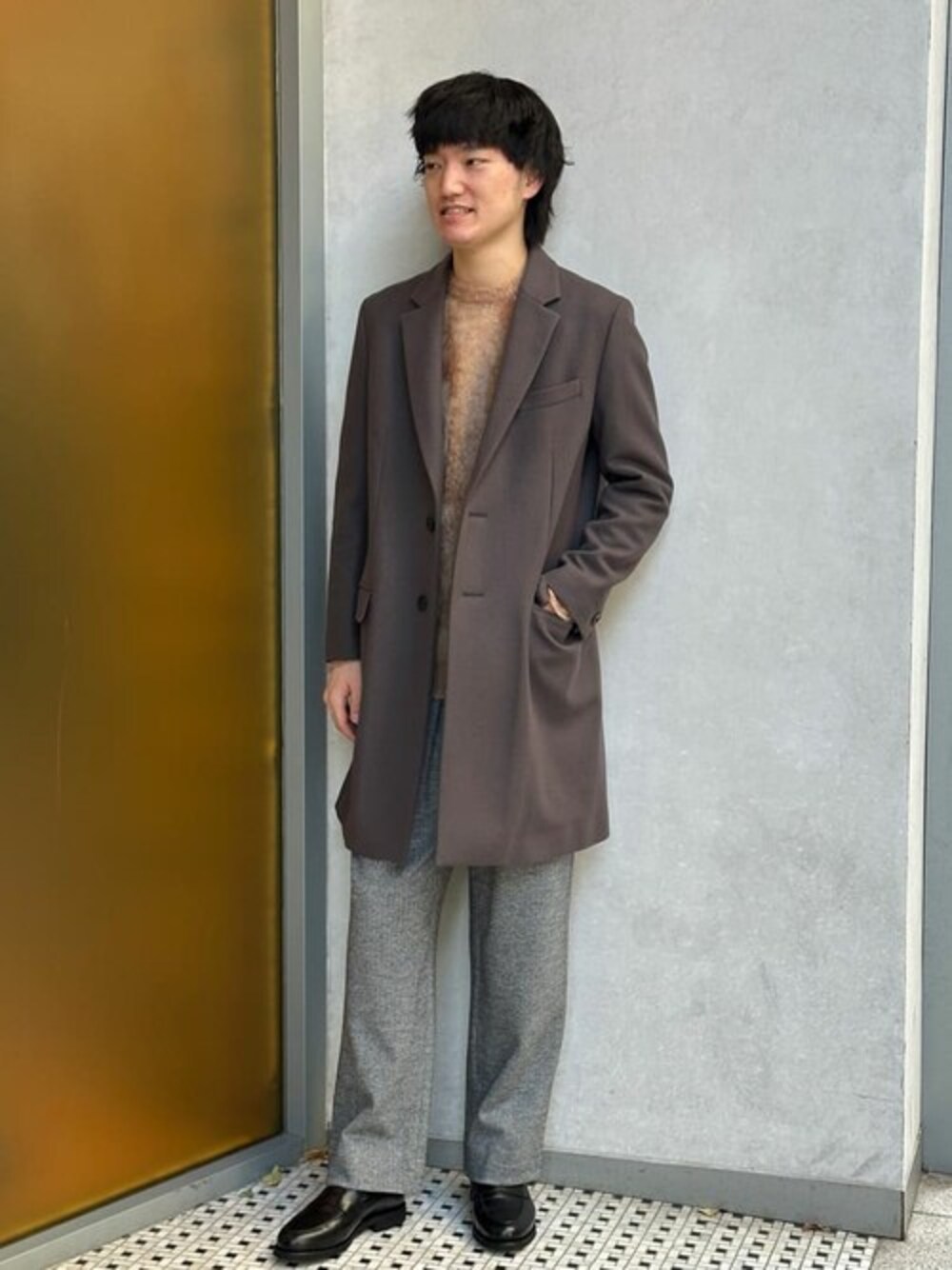 グレー系のチェスターコート、ブラウン系のニット/セーター、グレー系のスラックスを着用したメンズの冬コーディネートの1枚目の写真