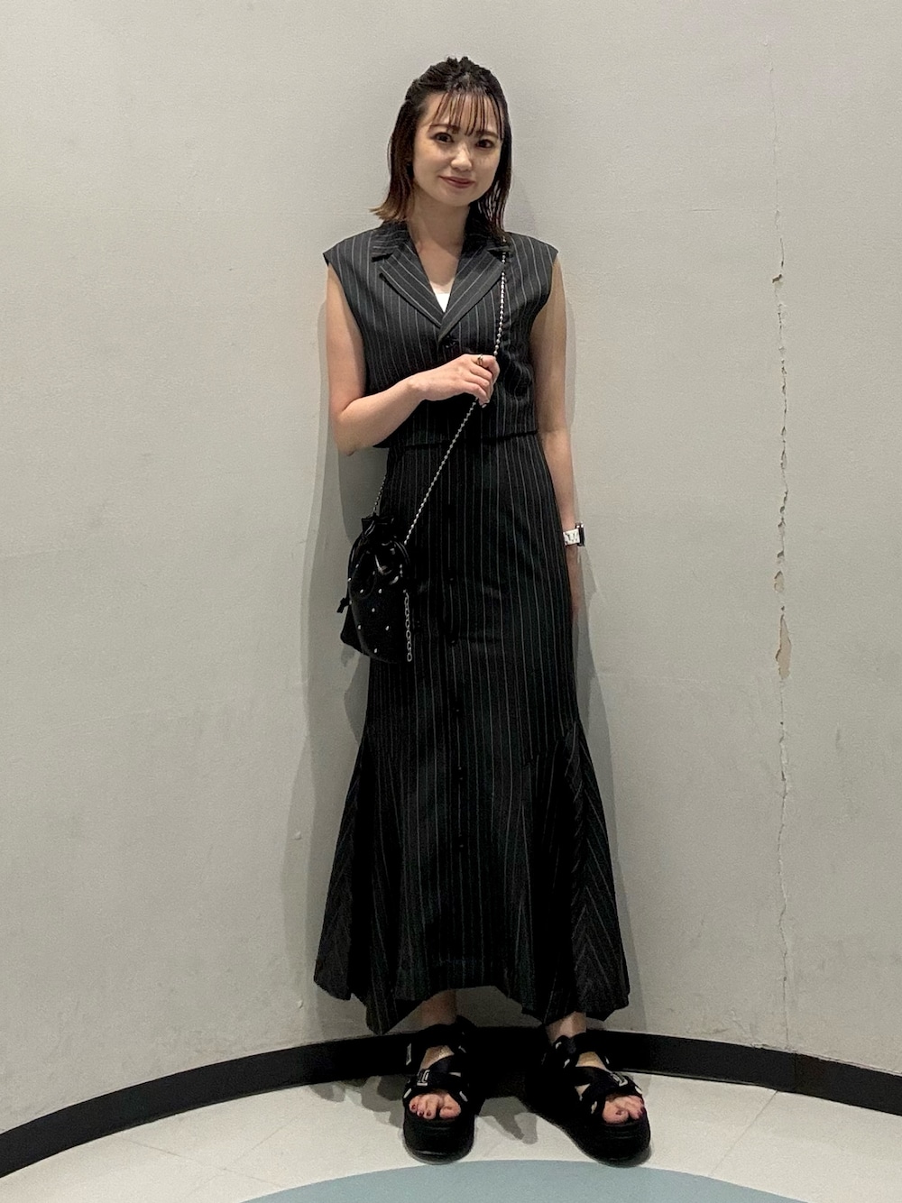 ブラック系のショルダーバッグ、ブラック系のサンダル、グレー系のジャンパースカートを着用したレディースの夏コーディネートの1枚目の写真