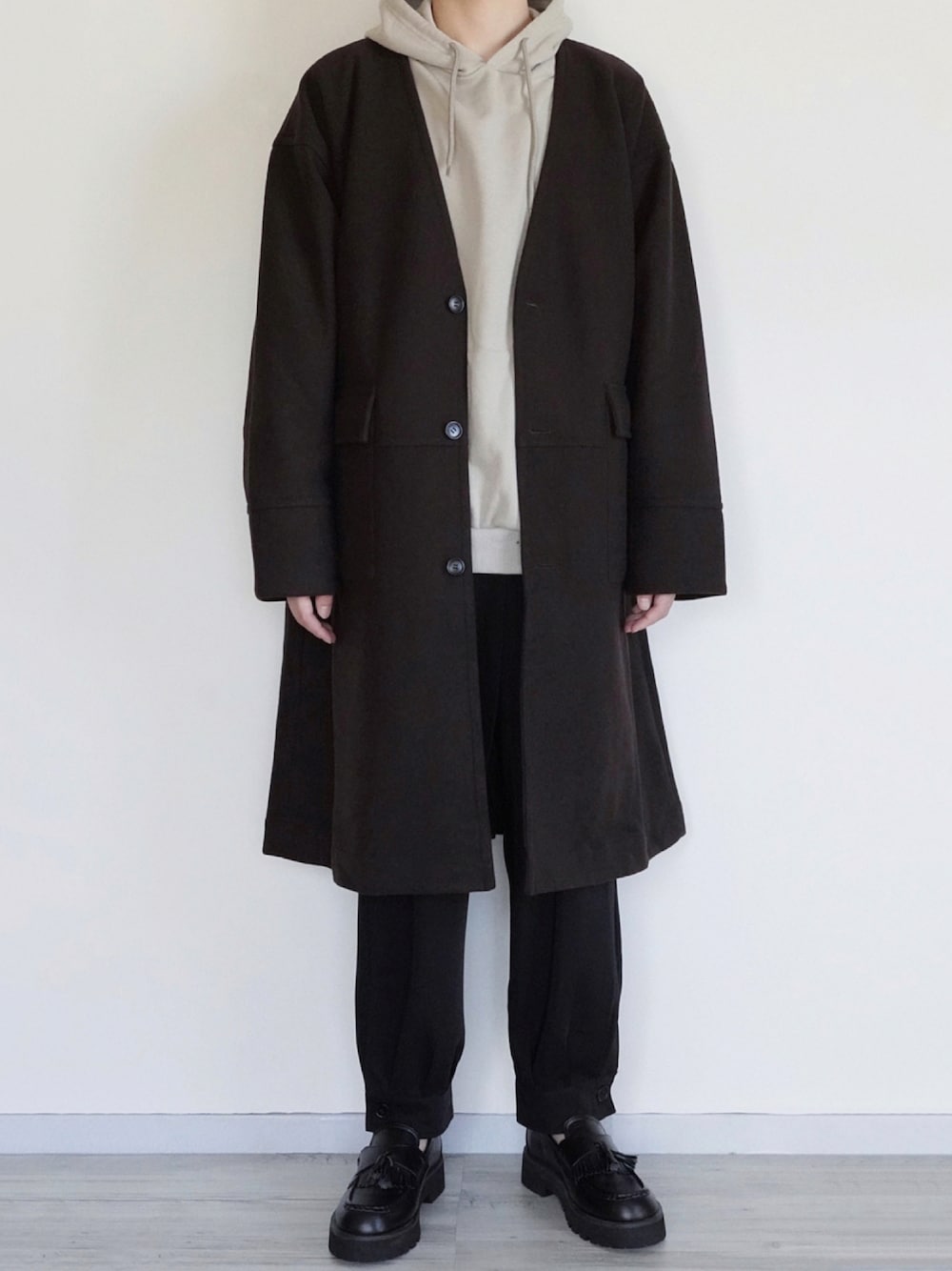 ブラウン系のノーカラーコート、グレー系のパーカー、ブラック系のその他パンツを着用したメンズの冬コーディネートの1枚目の写真