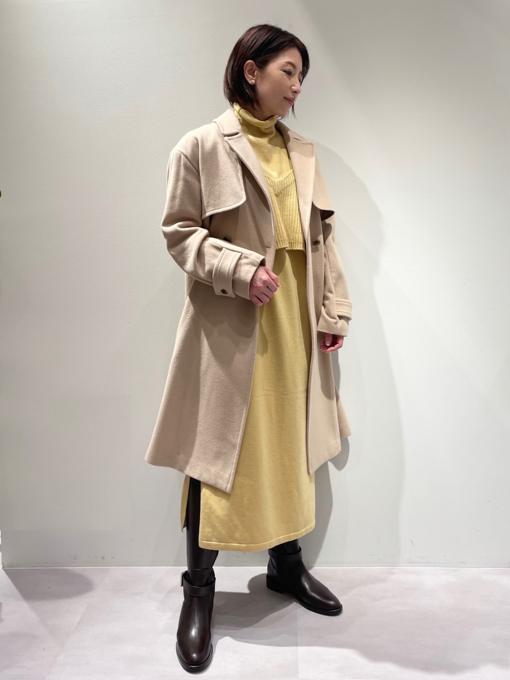 イエロー系のワンピース、ベージュ系のトレンチコート、ブラウン系のブーツを着用したレディースの冬コーディネートの1枚目の写真
