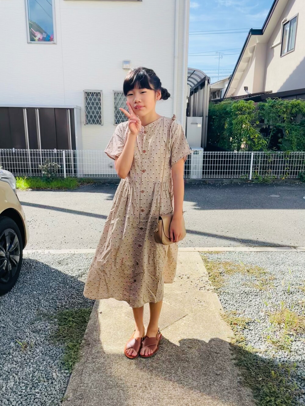 ベージュ系のシャツワンピース、ブラウン系のサンダル、ベージュ系のショルダーバッグを着用したキッズの夏コーディネートの1枚目の写真