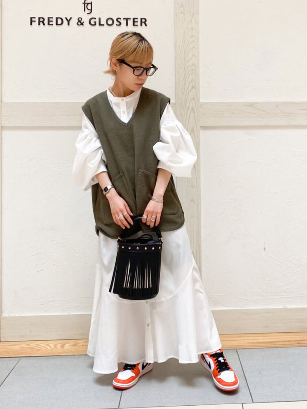 グリーン系のベスト、ホワイト系のシャツワンピース、ブラック系のショルダーバッグを着用したレディースの春コーディネートの1枚目の写真