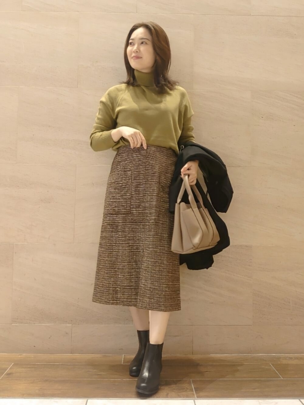 イエロー系のニット/セーター、ブラウン系のスカート、ブラック系のブーツを着用したレディースの秋コーディネートの1枚目の写真