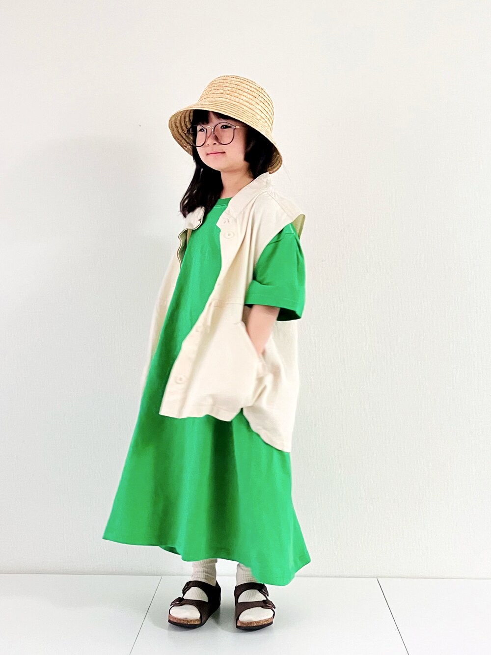 ホワイト系のベスト、グリーン系のワンピース、ブラウン系のサンダルを着用したキッズの春コーディネートの1枚目の写真
