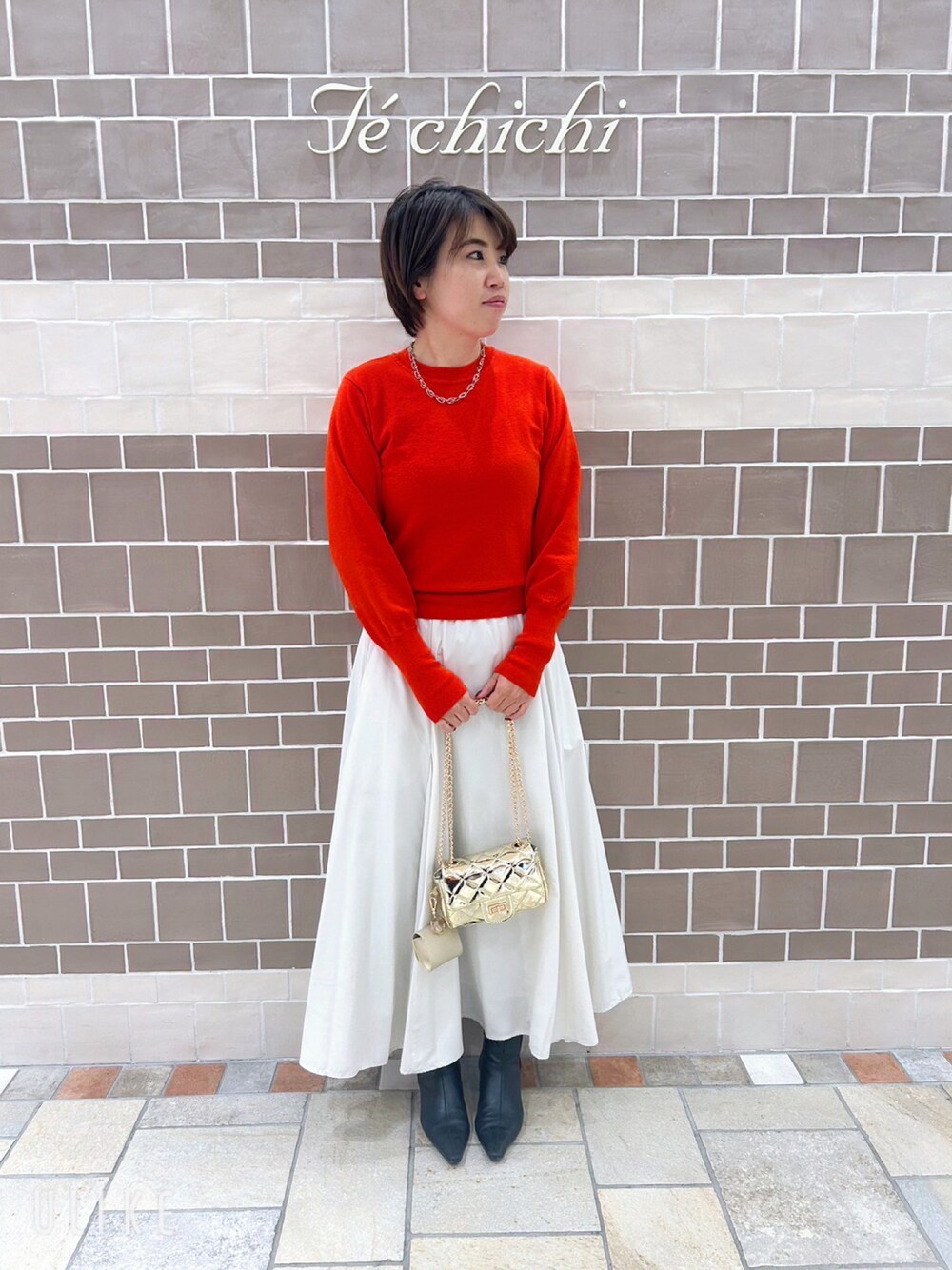 レッド系のニット/セーター、ゴールド系のハンドバッグ、ホワイト系のスカートを着用したレディースの冬コーディネートの1枚目の写真