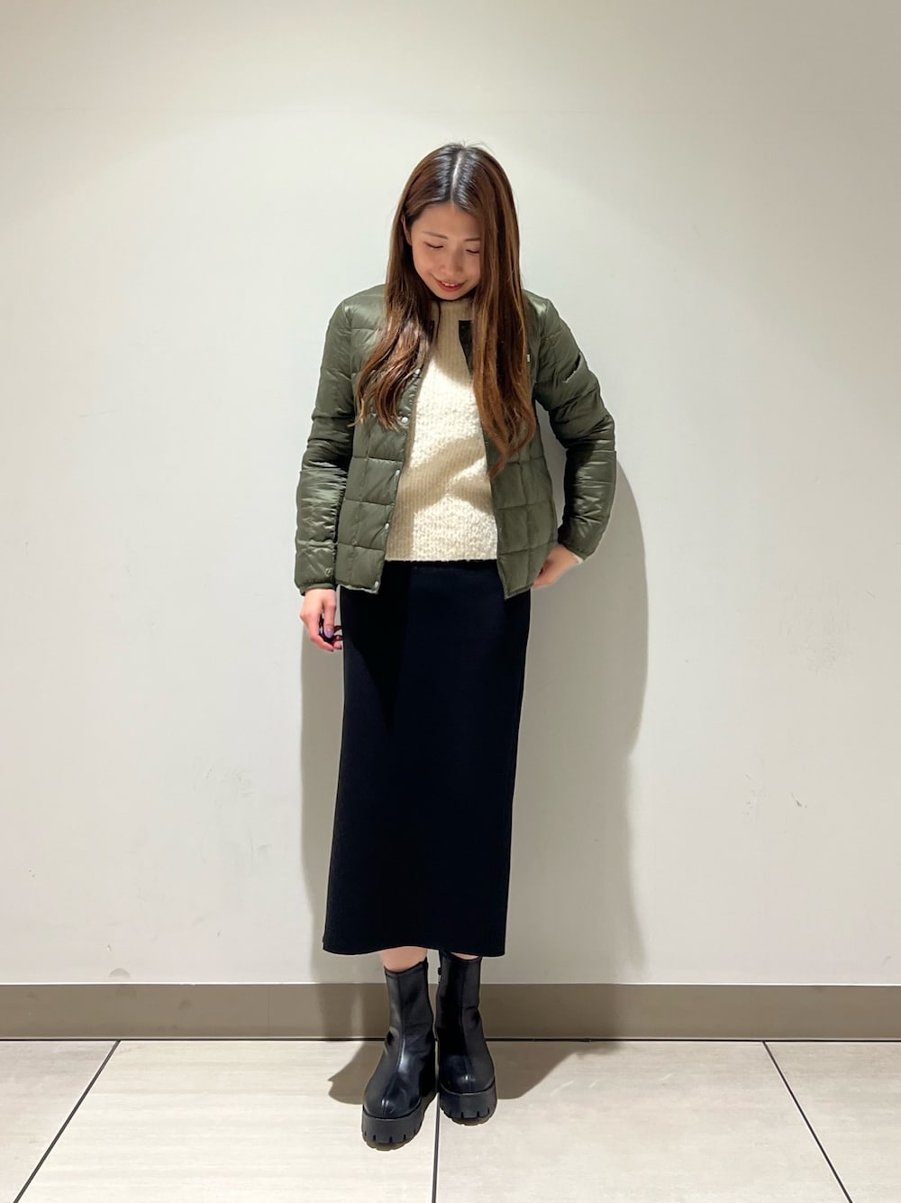 ブラック系のスカート、グリーン系のダウンジャケット/コート、ホワイト系のニット/セーターを着用したレディースの冬コーディネートの1枚目の写真