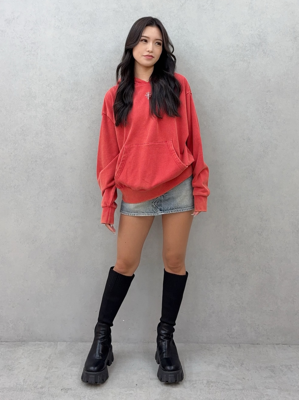 レッド系のパーカー、ブルー系のデニムスカート、ブラック系のブーツを着用したレディースの秋コーディネートの1枚目の写真