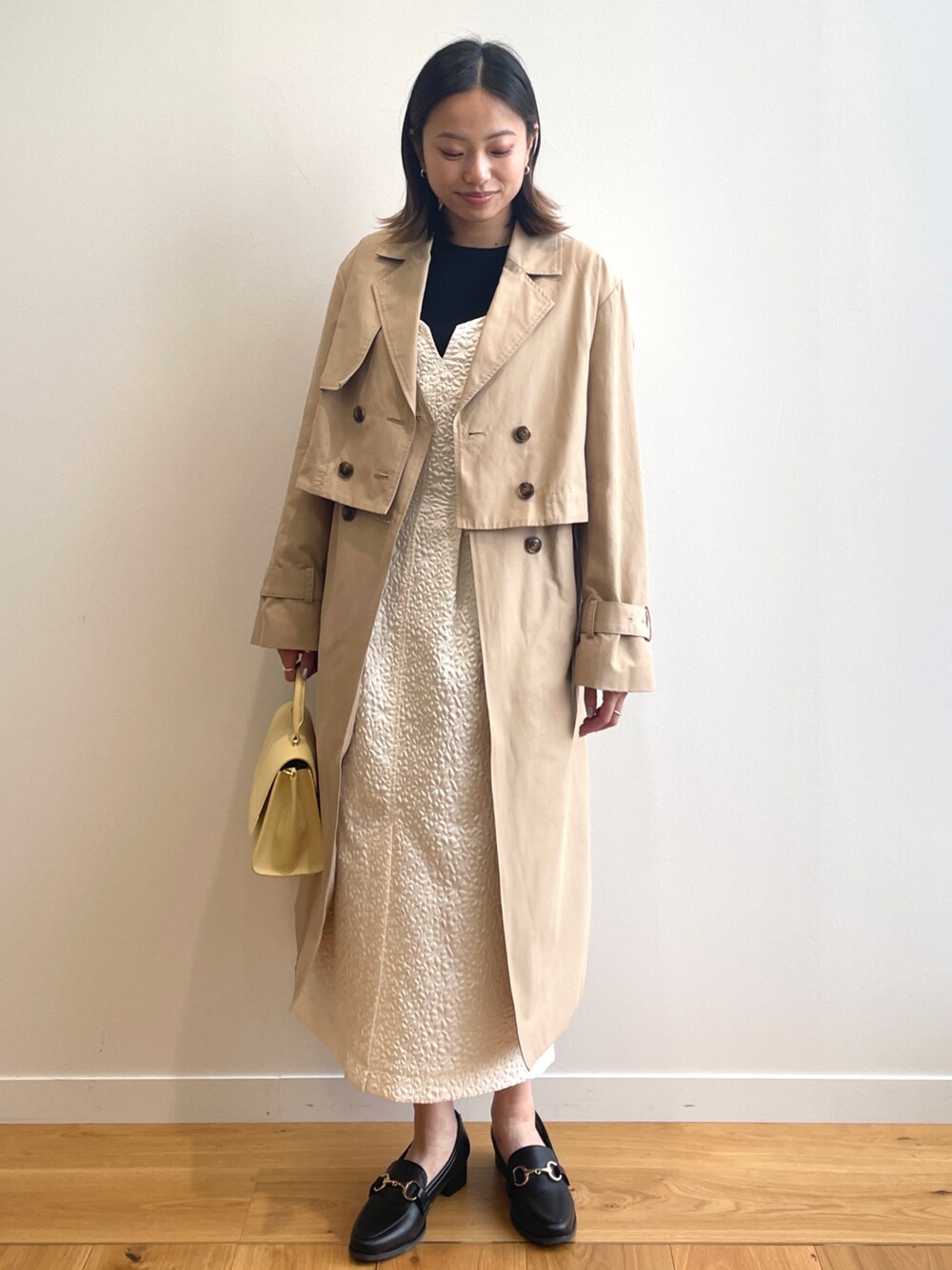 ベージュ系のトレンチコート、ホワイト系のワンピース、イエロー系のショルダーバッグを着用したレディースの春コーディネートの1枚目の写真