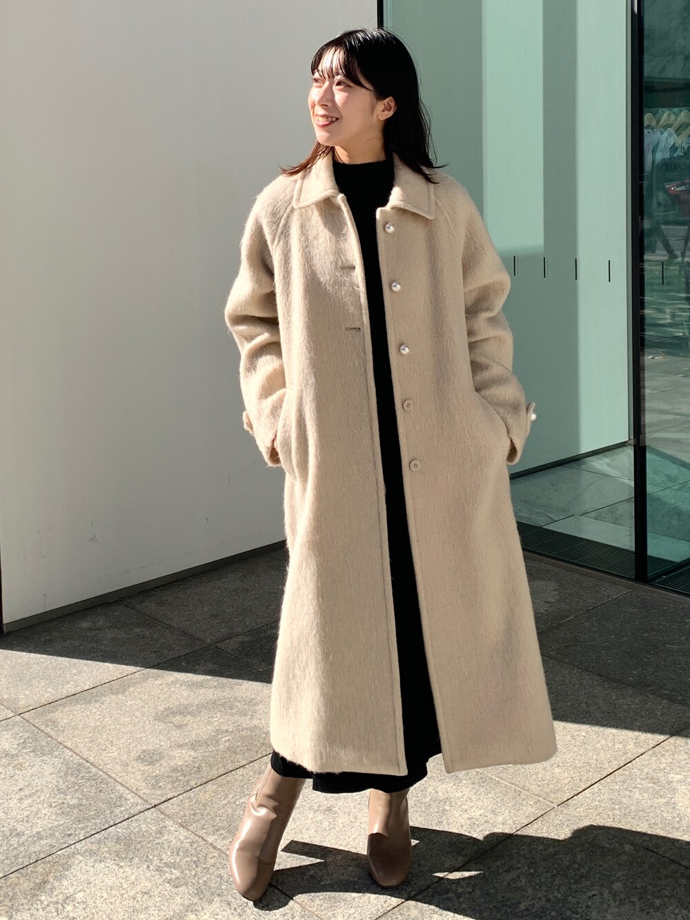 ベージュ系のステンカラーコート、ブラック系のワンピース、ブラウン系のブーツを着用したレディースの秋コーディネートの1枚目の写真