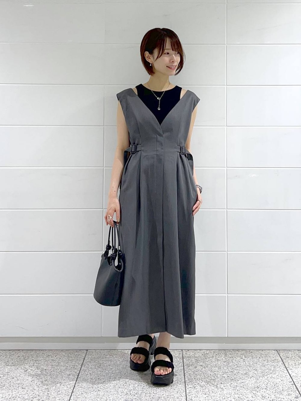 ブラック系のハンドバッグ、ブラック系のサンダル、グレー系のジャンパースカートを着用したレディースの夏コーディネートの1枚目の写真