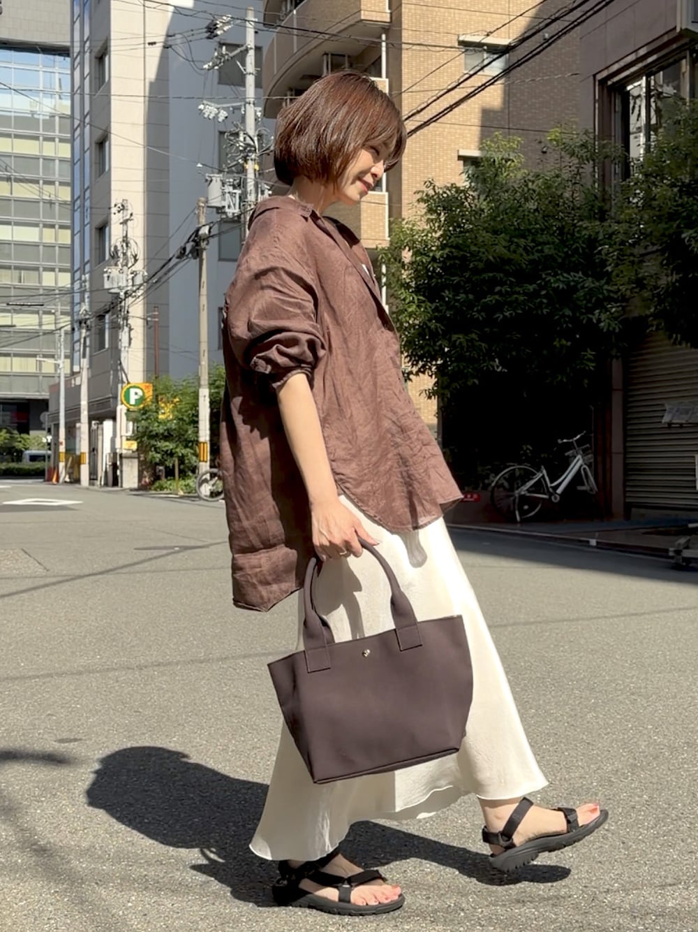 グレー系のトートバッグ、ブラウン系のシャツ/ブラウス、ブラック系のサンダルを着用したレディースの春コーディネートの1枚目の写真