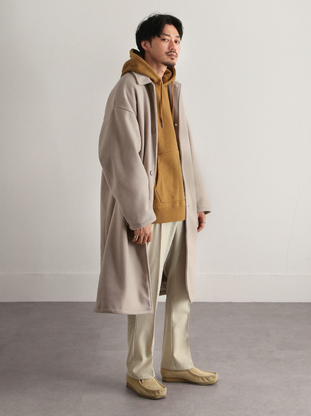 ベージュ系のステンカラーコート、イエロー系のパーカー、ベージュ系のスラックスを着用したメンズの冬コーディネートの1枚目の写真