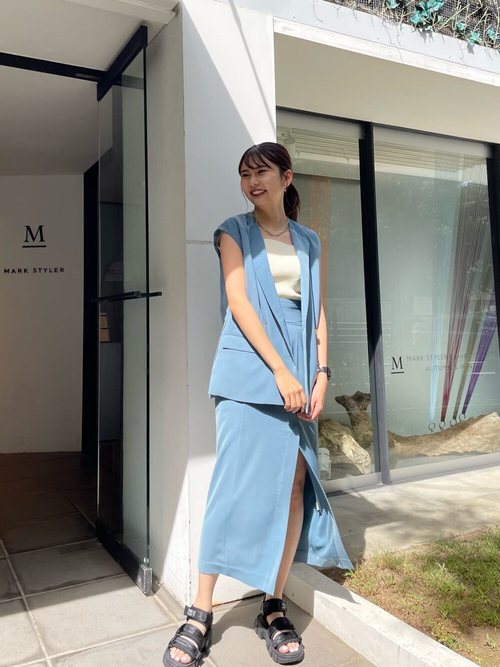 ブルー系のスカート、ブルー系のベスト、ブラック系のサンダルを着用したレディースの夏コーディネートの1枚目の写真