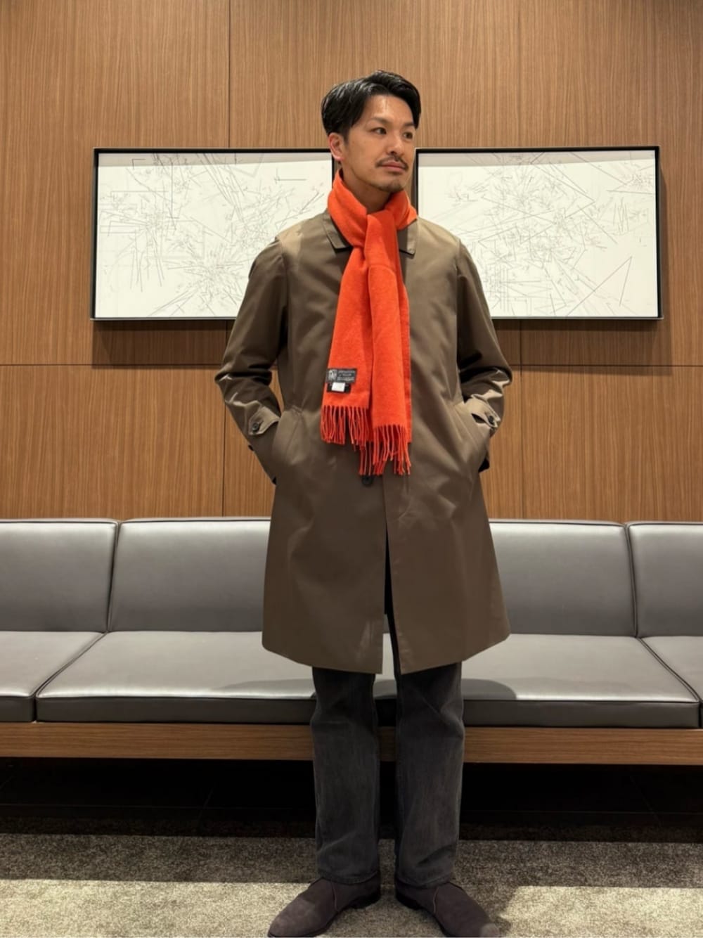 グリーン系のステンカラーコート、グレー系のニット/セーター、オレンジ系のマフラーを着用したメンズの冬コーディネートの1枚目の写真