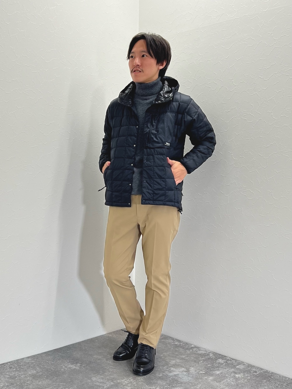 グレー系のニット/セーター、ブラック系のダウンジャケット/コート、ベージュ系のチノパンツを着用したメンズの秋コーディネートの1枚目の写真