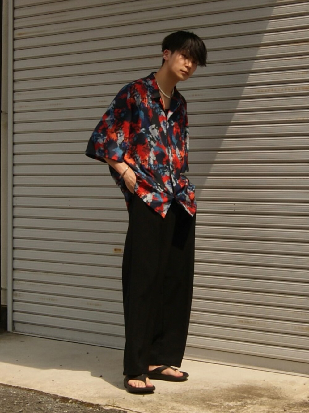 レッド系のトップス、ブラック系のスラックス、ブラック系のサンダルを着用したメンズの春コーディネートの1枚目の写真