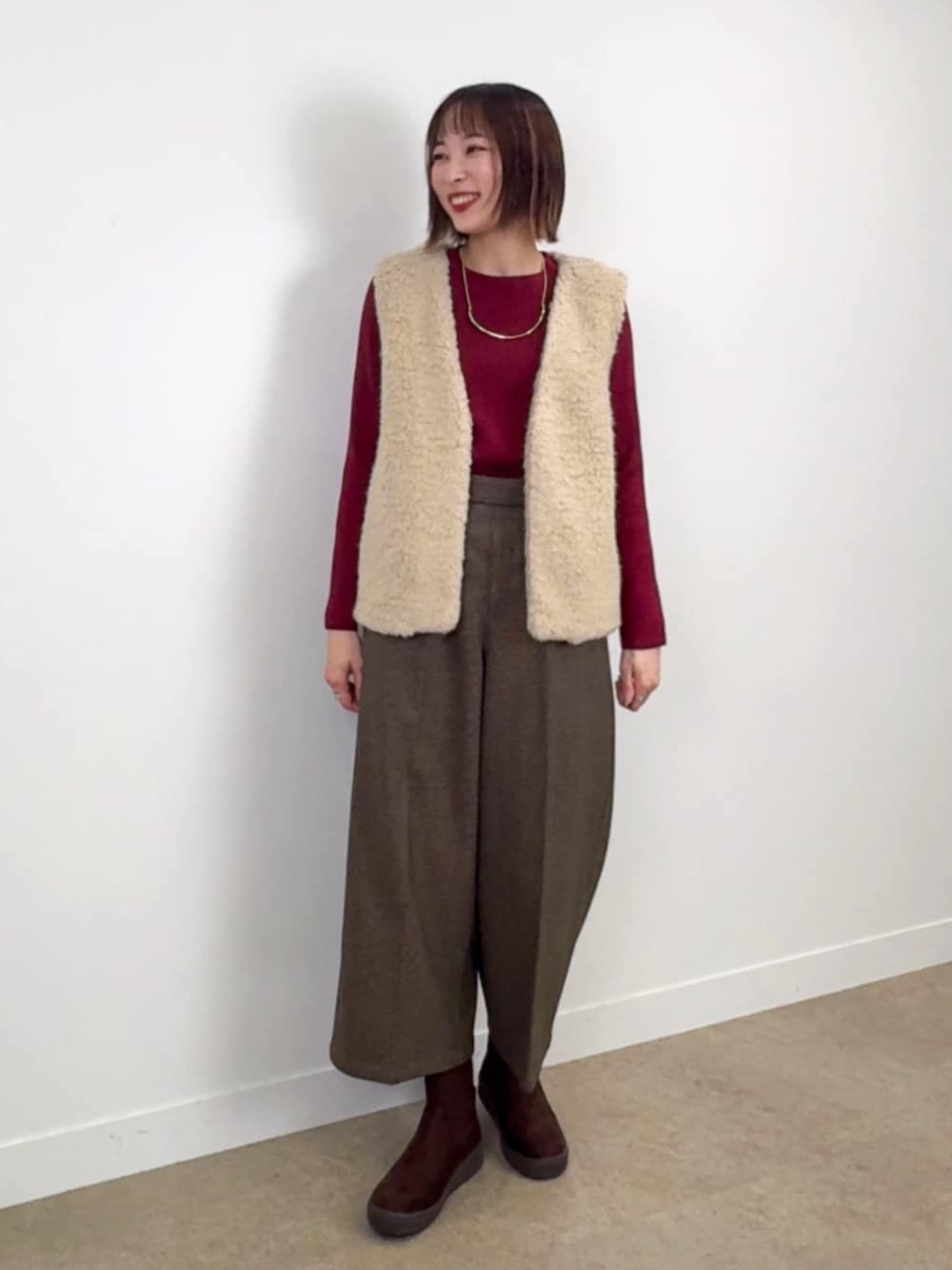 レッド系のニット/セーター、ベージュ系のベスト、ブラウン系のブーツを着用したレディースの秋コーディネートの1枚目の写真