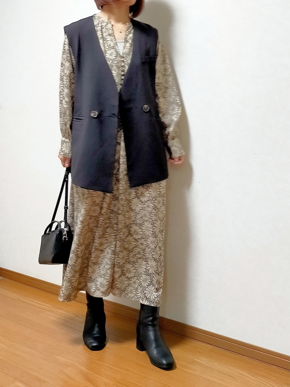 ブラック系のベスト、ベージュ系のワンピース、ブラック系のブーツを着用したレディースの春コーディネートの2枚目の写真