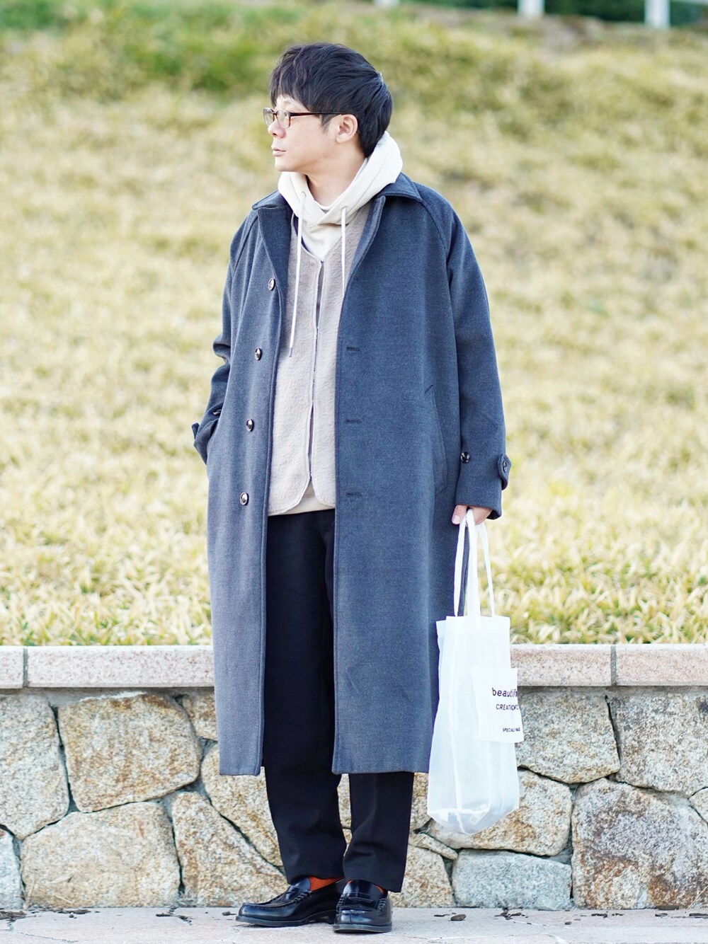 グレー系のステンカラーコート、ベージュ系のパーカー、ベージュ系のベストを着用したメンズの冬コーディネートの1枚目の写真