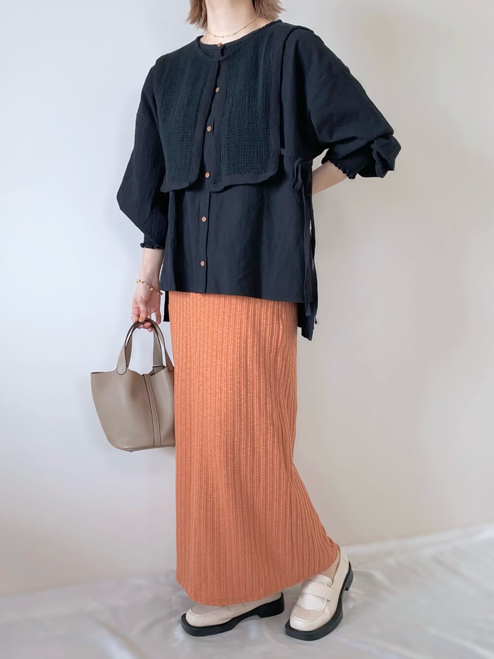 オレンジ系のスカート、ベージュ系のローファー、ブラック系のシャツ/ブラウスを着用したレディースの春コーディネートの1枚目の写真