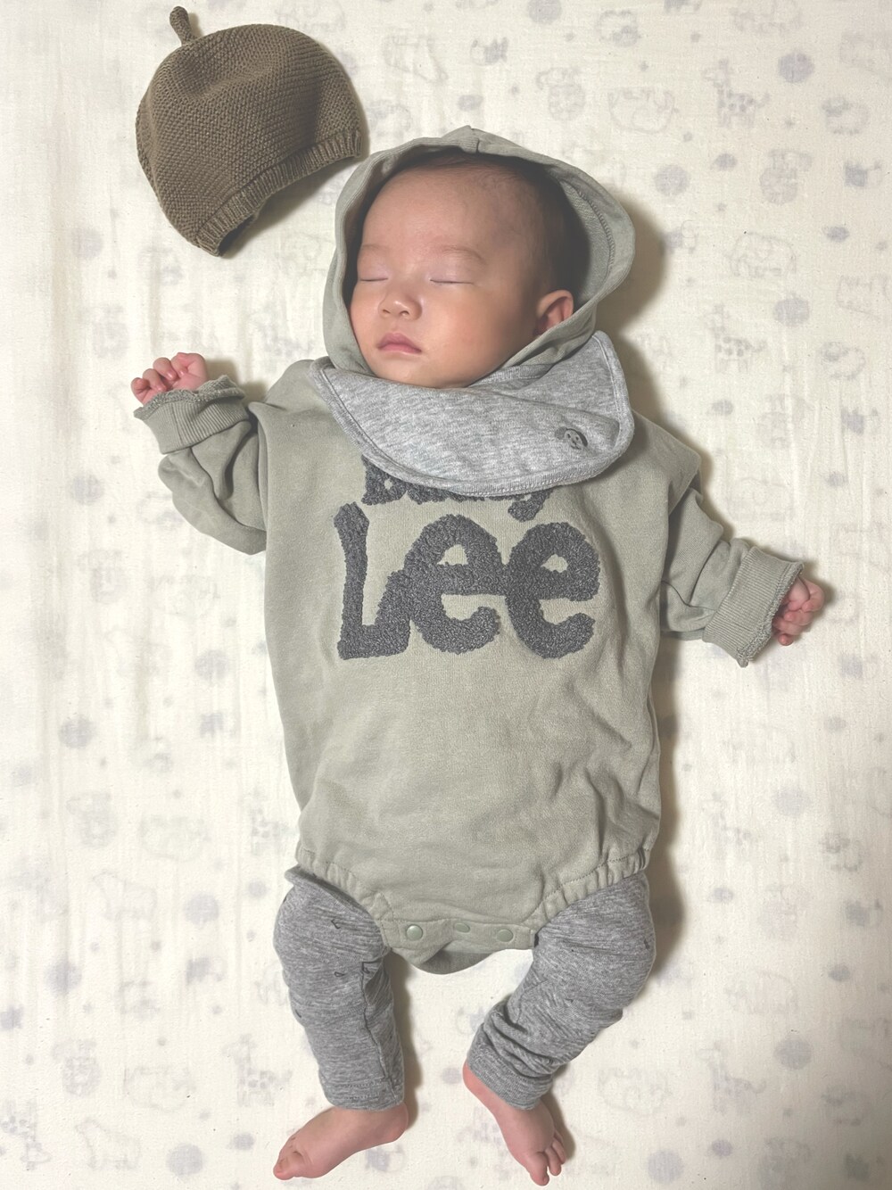 グリーン系のロンパース、グレー系のスタイ/よだれかけ、グリーン系の帽子を着用したキッズの冬コーディネートの1枚目の写真