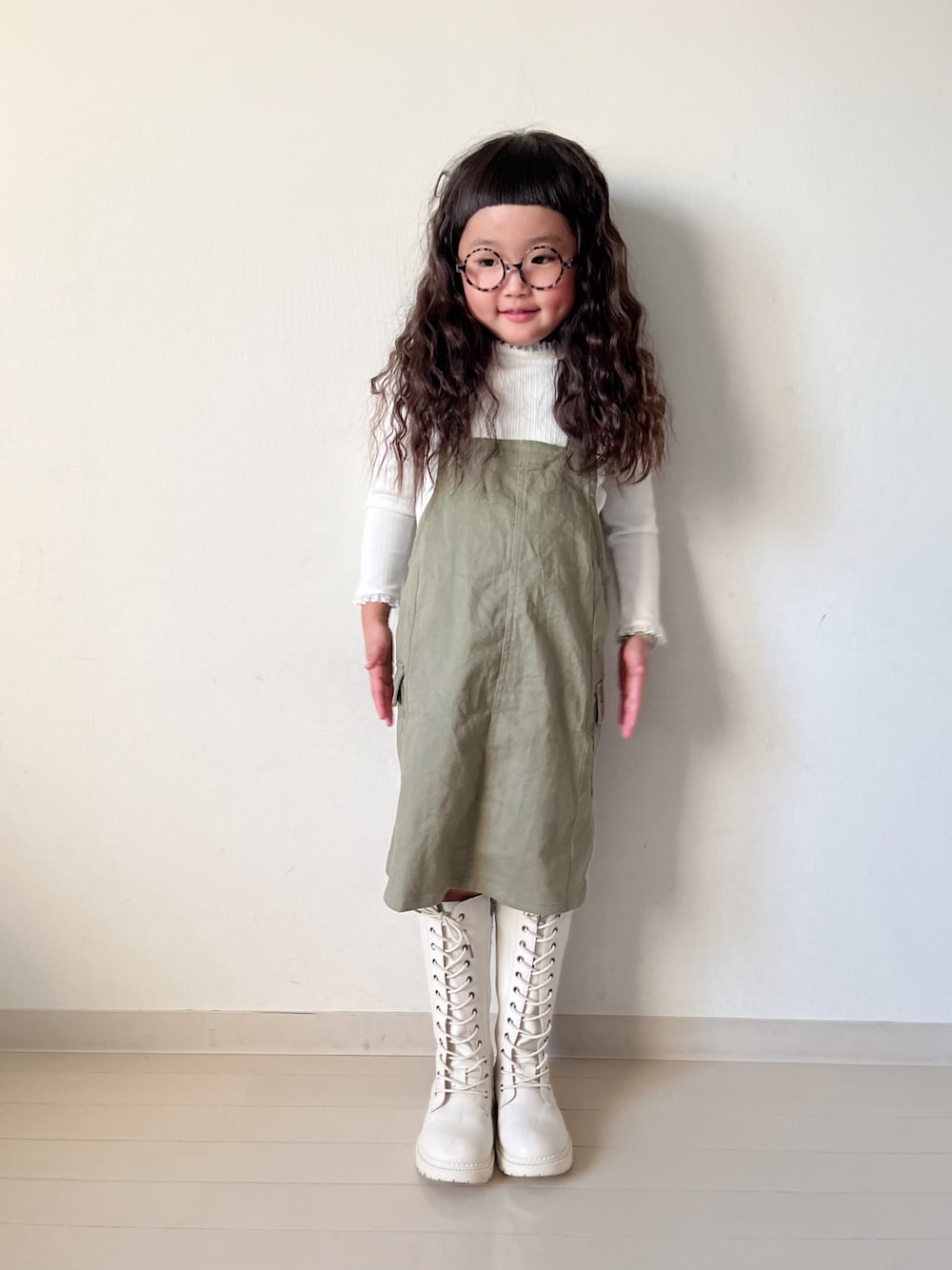 グリーン系のジャンパースカート、ベージュ系のTシャツ/カットソー、ホワイト系のカーディガン/ボレロを着用したキッズの冬コーディネートの1枚目の写真