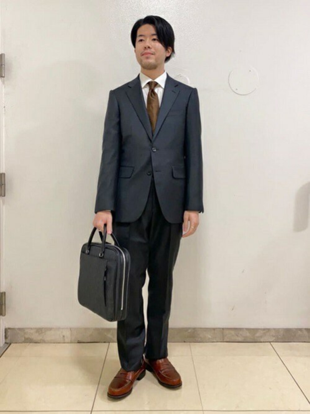 グレー系のスーツセット、ブラウン系のネクタイ、ブラック系のビジネスバッグを着用したメンズの春コーディネートの1枚目の写真