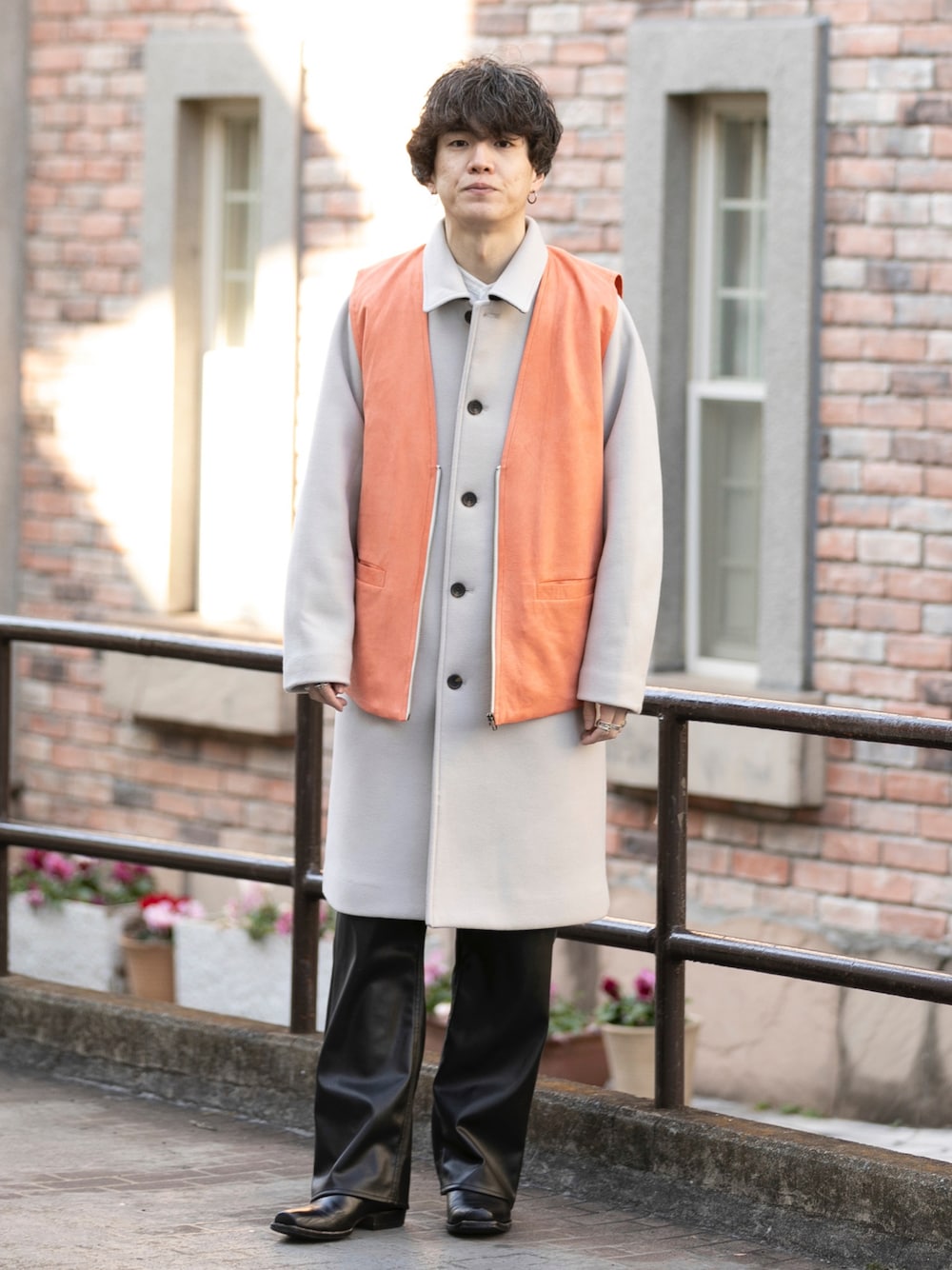 ベージュ系のステンカラーコート、オレンジ系のベスト、ブラック系のその他パンツを着用したメンズの冬コーディネートの1枚目の写真