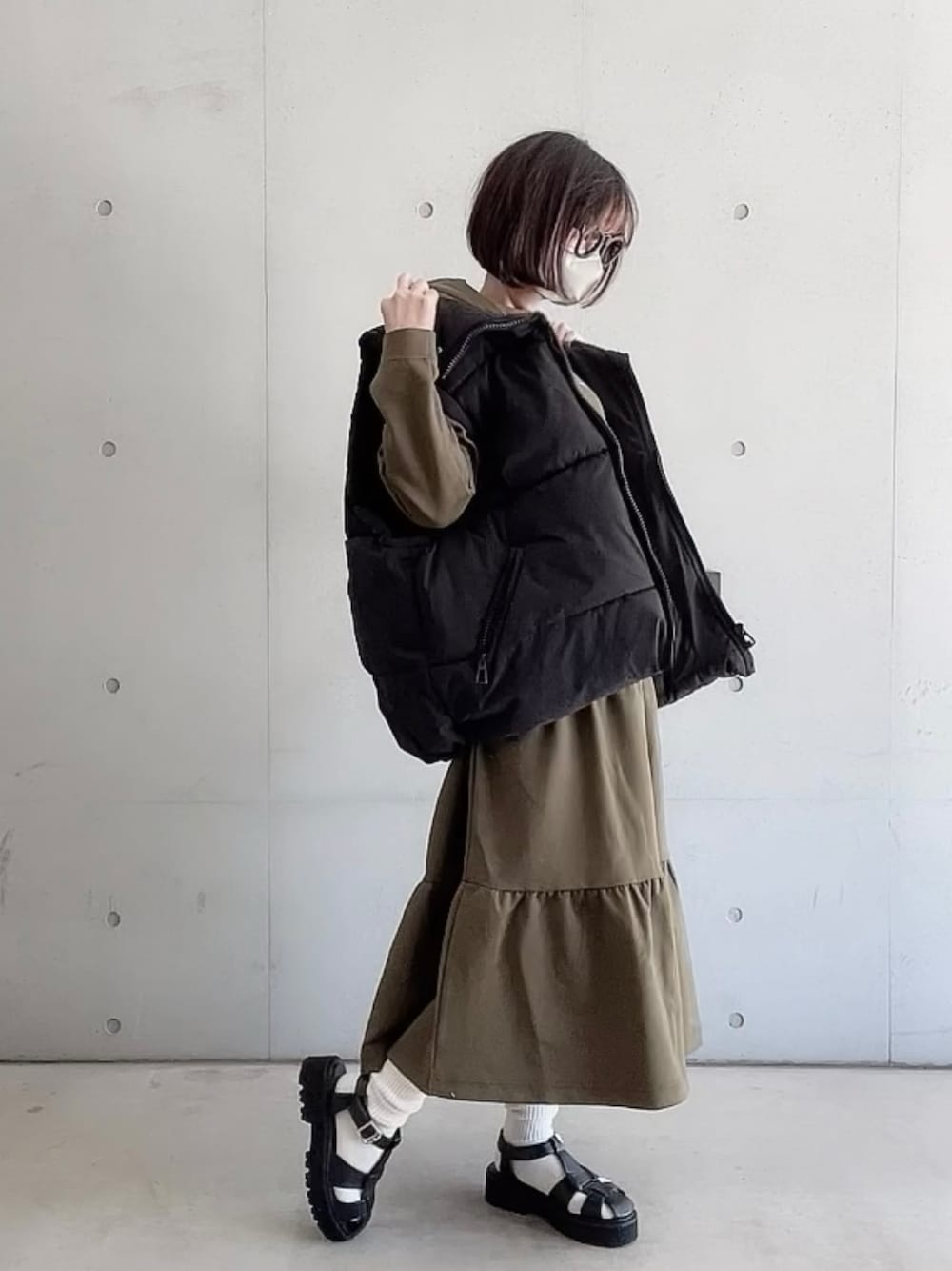 グリーン系のワンピース、ブラック系のダウンベスト、ブラック系のサンダルを着用したレディースの春コーディネートの1枚目の写真