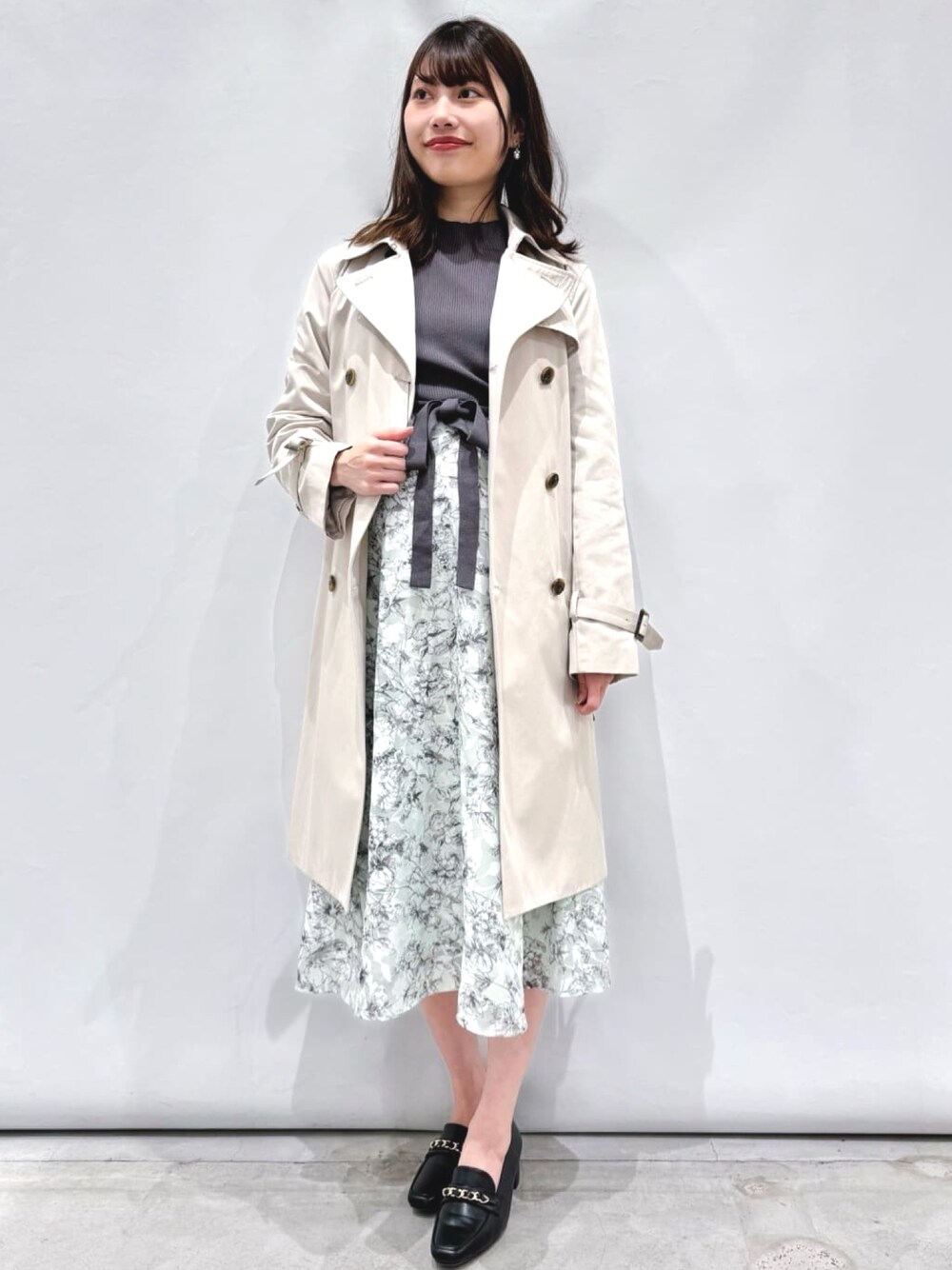 グレー系のワンピース、ベージュ系のトレンチコート、ブラック系のパンプスを着用したレディースの春コーディネートの1枚目の写真