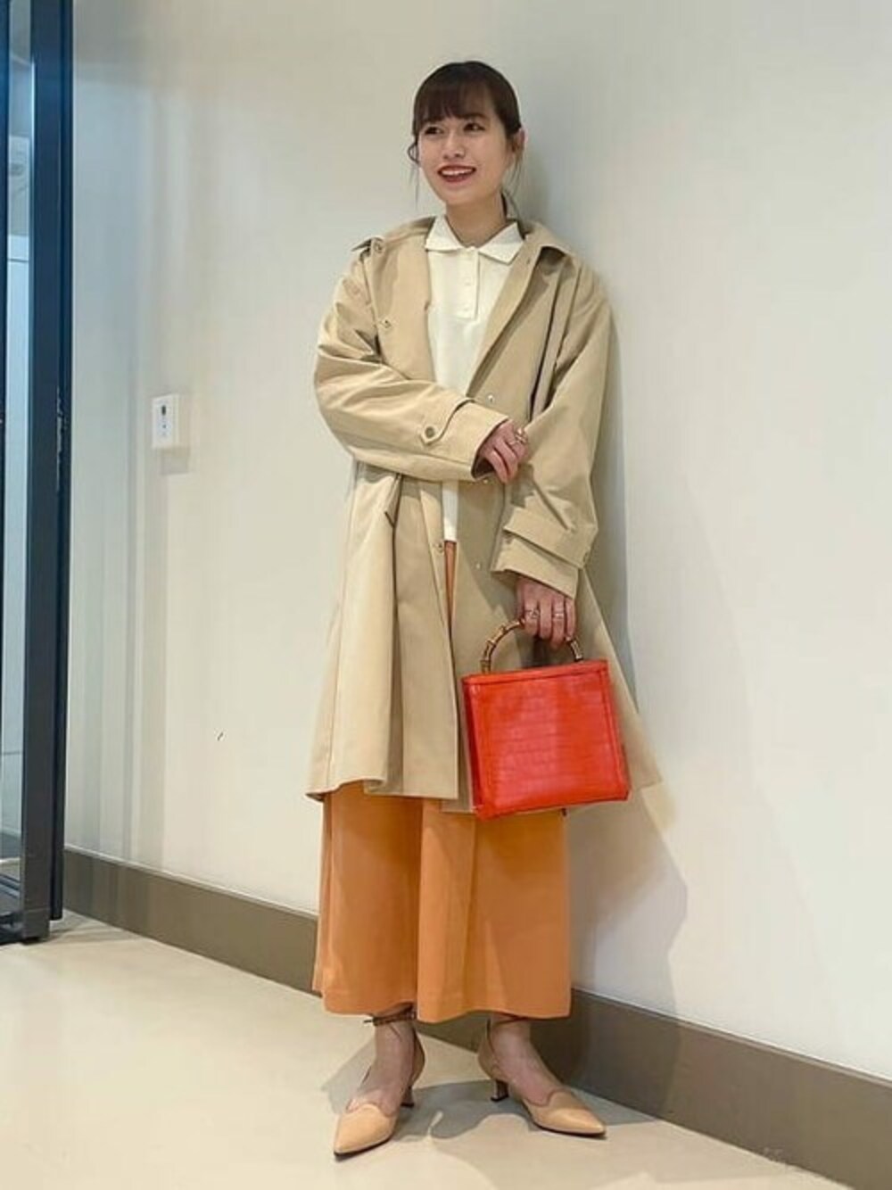 ベージュ系のステンカラーコート、ホワイト系のポロシャツ、オレンジ系のスカートを着用したレディースの冬コーディネートの1枚目の写真