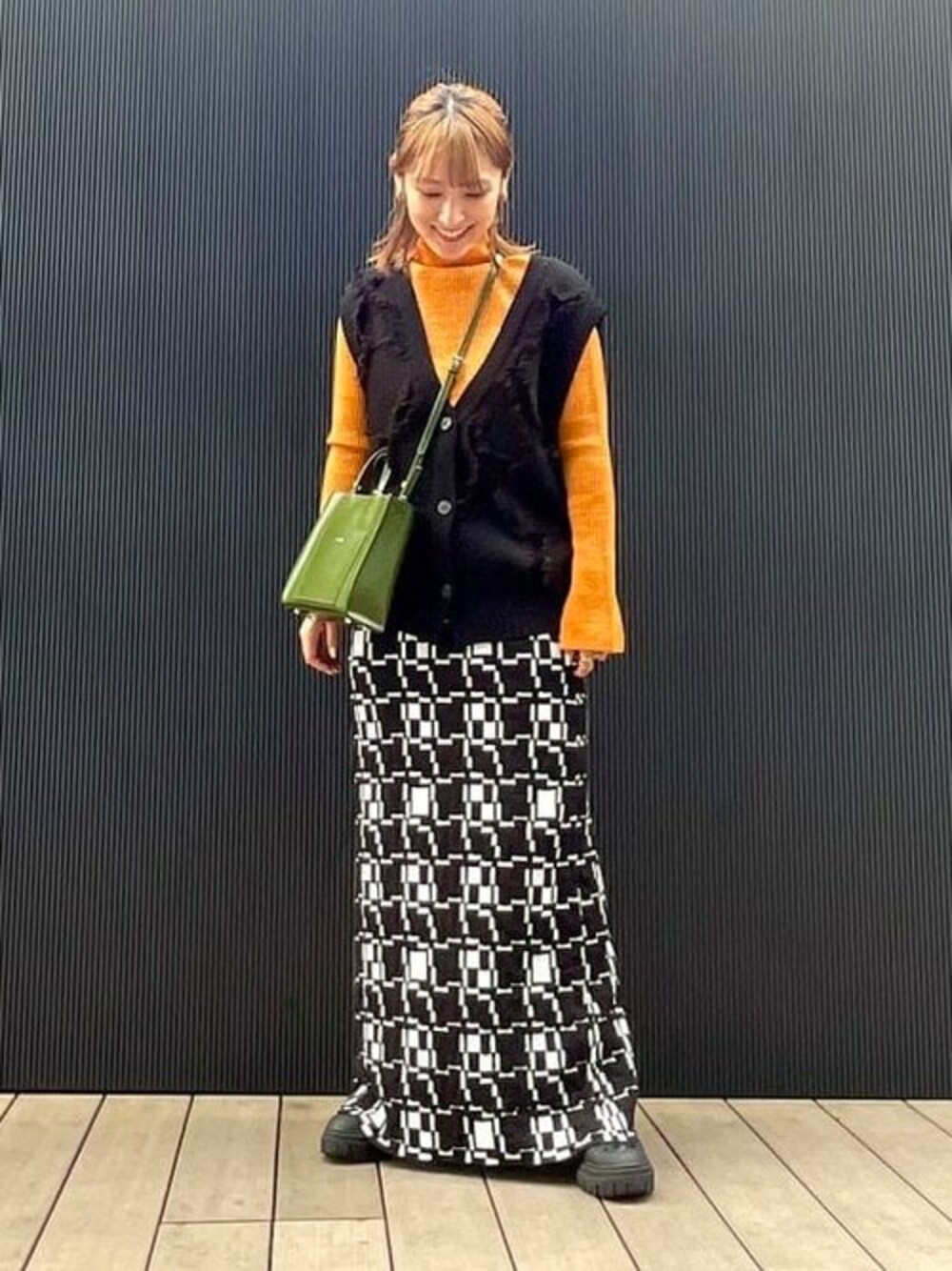 ブラック系のベスト、オレンジ系のニット/セーター、ブラック系のスカートを着用したレディースの秋コーディネートの1枚目の写真