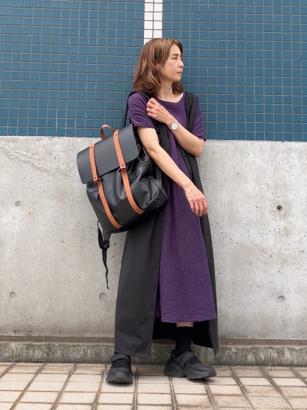 ブラック系のジャンパースカート、パープル系のワンピース、ブラウン系のバックパック/リュックを着用したレディースの春コーディネートの1枚目の写真