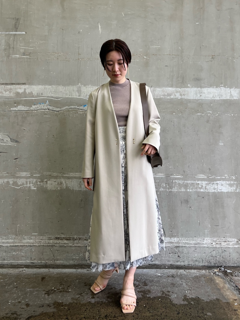 ベージュ系のノーカラーコート、ホワイト系のスカート、ベージュ系のサンダルを着用したレディースの冬コーディネートの1枚目の写真