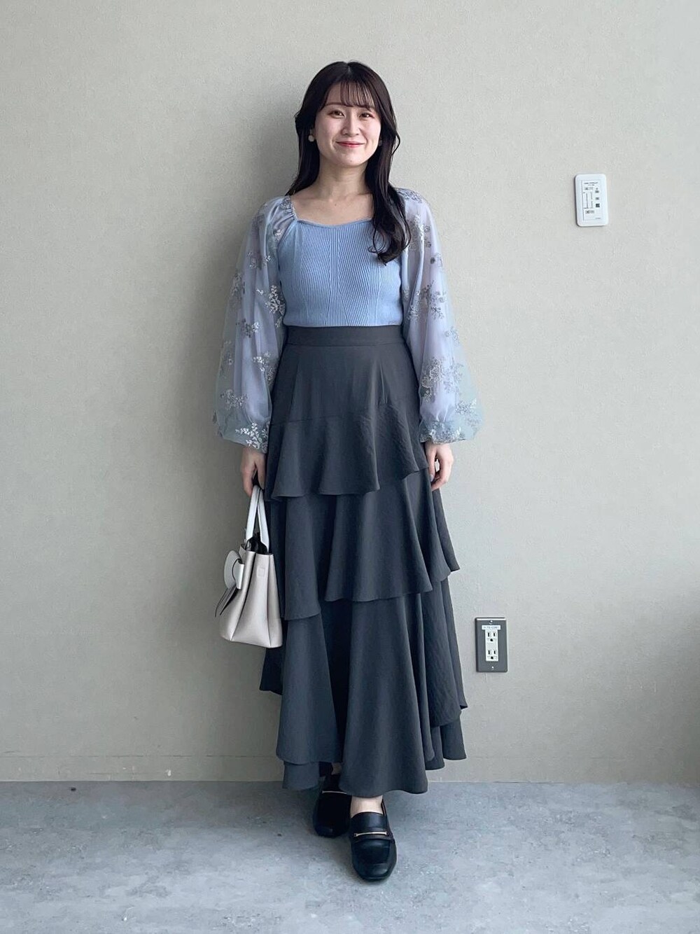 グレー系のスカート、ベージュ系のショルダーバッグ、ブルー系のニット/セーターを着用したレディースの春コーディネートの1枚目の写真