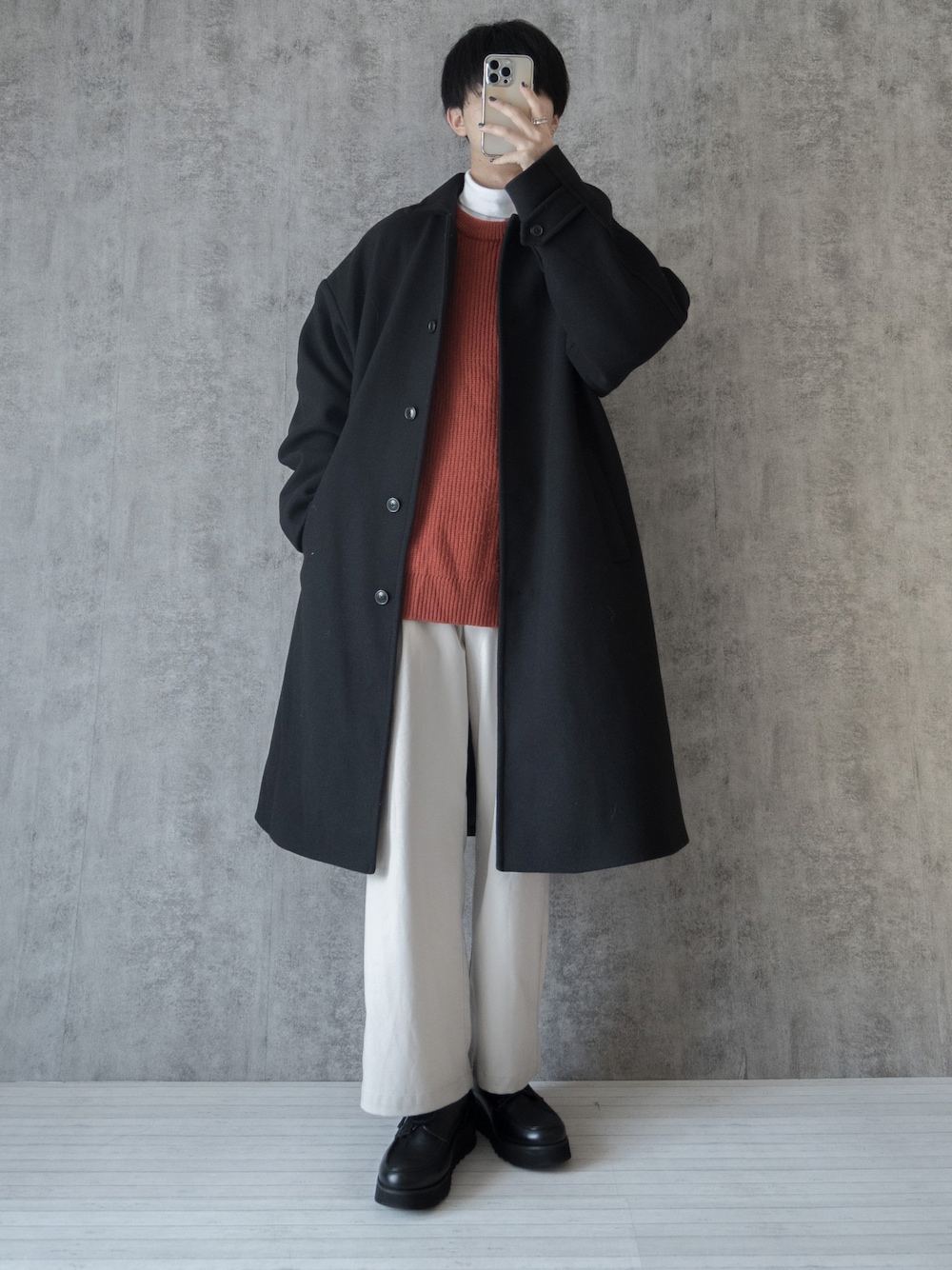 ブラック系のステンカラーコート、オレンジ系のニット/セーター、ホワイト系のTシャツ/カットソーを着用したメンズの冬コーディネートの1枚目の写真
