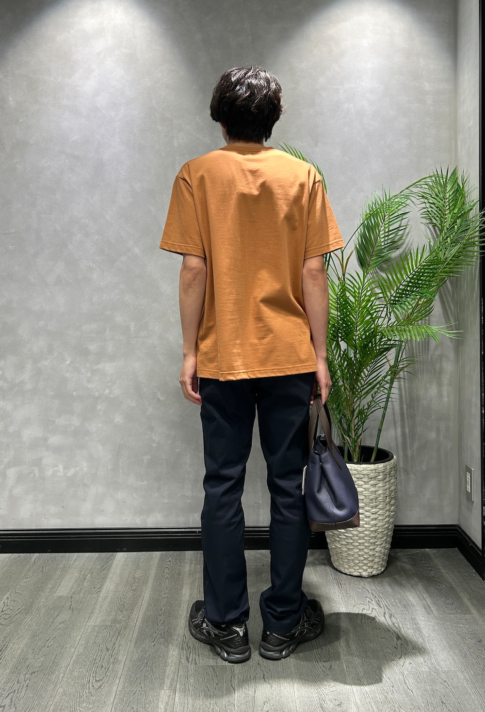 オレンジ系のTシャツ/カットソー、ブルー系のスラックス、ブルー系のトートバッグを着用したメンズの夏コーディネートの3枚目の写真
