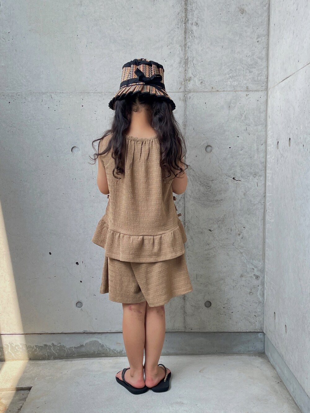 ブラウン系のパンツドレス、ブラック系のサンダル、ブラック系の帽子を着用したキッズの夏コーディネートの4枚目の写真