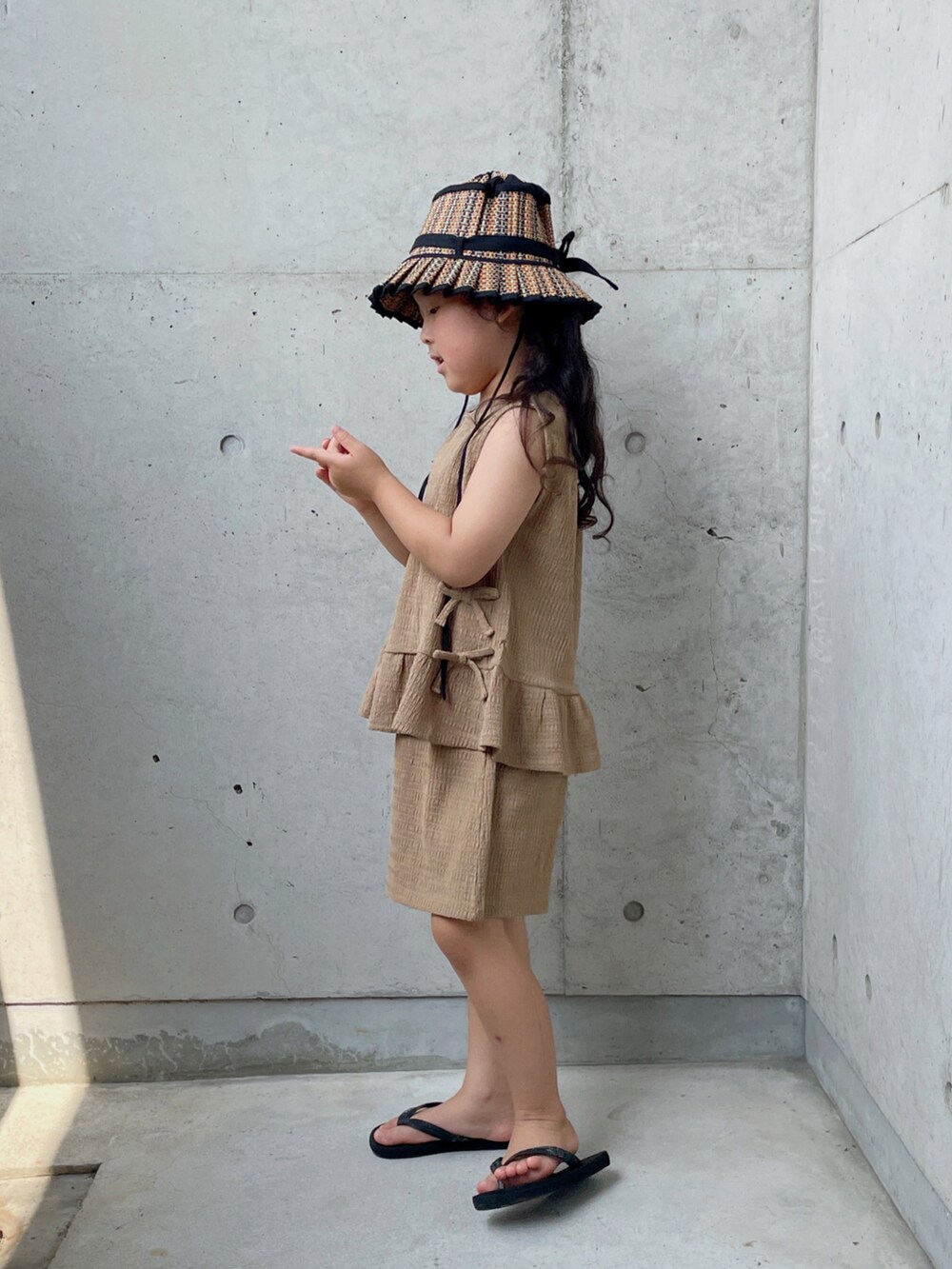 ブラウン系のパンツドレス、ブラック系のサンダル、ブラック系の帽子を着用したキッズの夏コーディネートの2枚目の写真