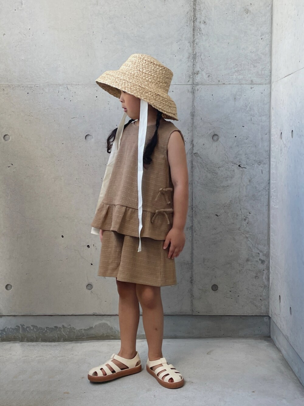 ブラウン系のパンツドレス、ホワイト系のサンダル、ホワイト系のハットを着用したキッズの夏コーディネートの2枚目の写真