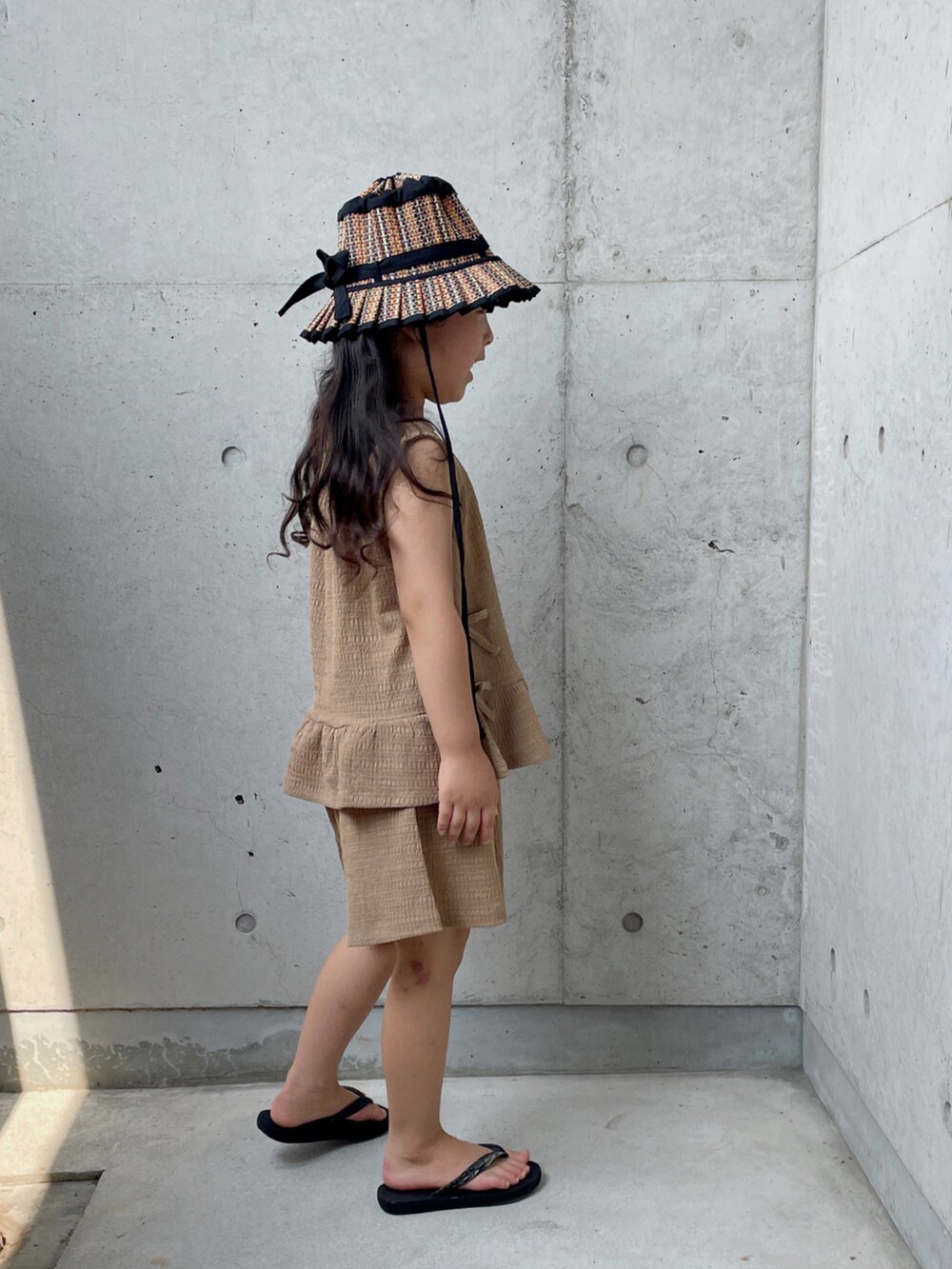 ブラウン系のパンツドレス、ブラック系のサンダル、ブラック系の帽子を着用したキッズの夏コーディネートの3枚目の写真