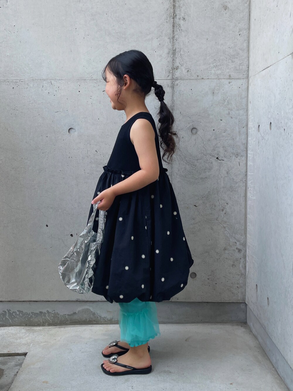 シルバー系のハンドバッグ、ブラック系のワンピース、ブラック系のサンダルを着用したキッズの夏コーディネートの2枚目の写真