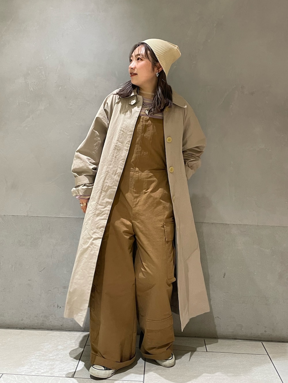 ベージュ系のステンカラーコート、イエロー系のつなぎ/オールインワン、パープル系のTシャツ/カットソーを着用したレディースの冬コーディネートの2枚目の写真