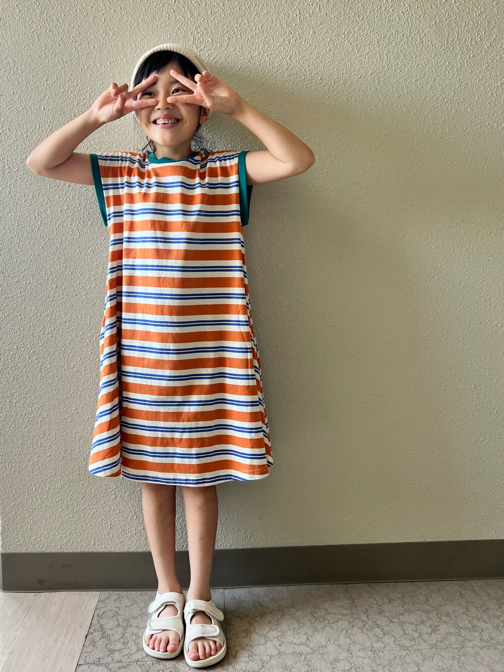オレンジ系のワンピース、ベージュ系のサンダル、ホワイト系のキャップを着用したキッズの夏コーディネートの2枚目の写真