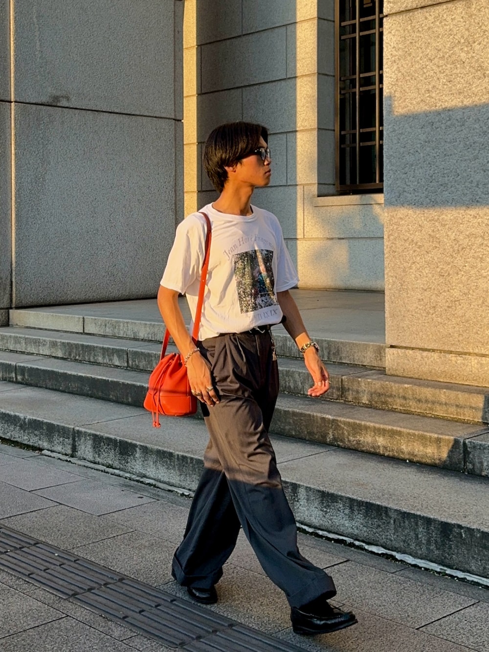 ホワイト系のTシャツ/カットソー、オレンジ系のバッグ、グレー系のスラックスを着用したメンズの夏コーディネートの3枚目の写真