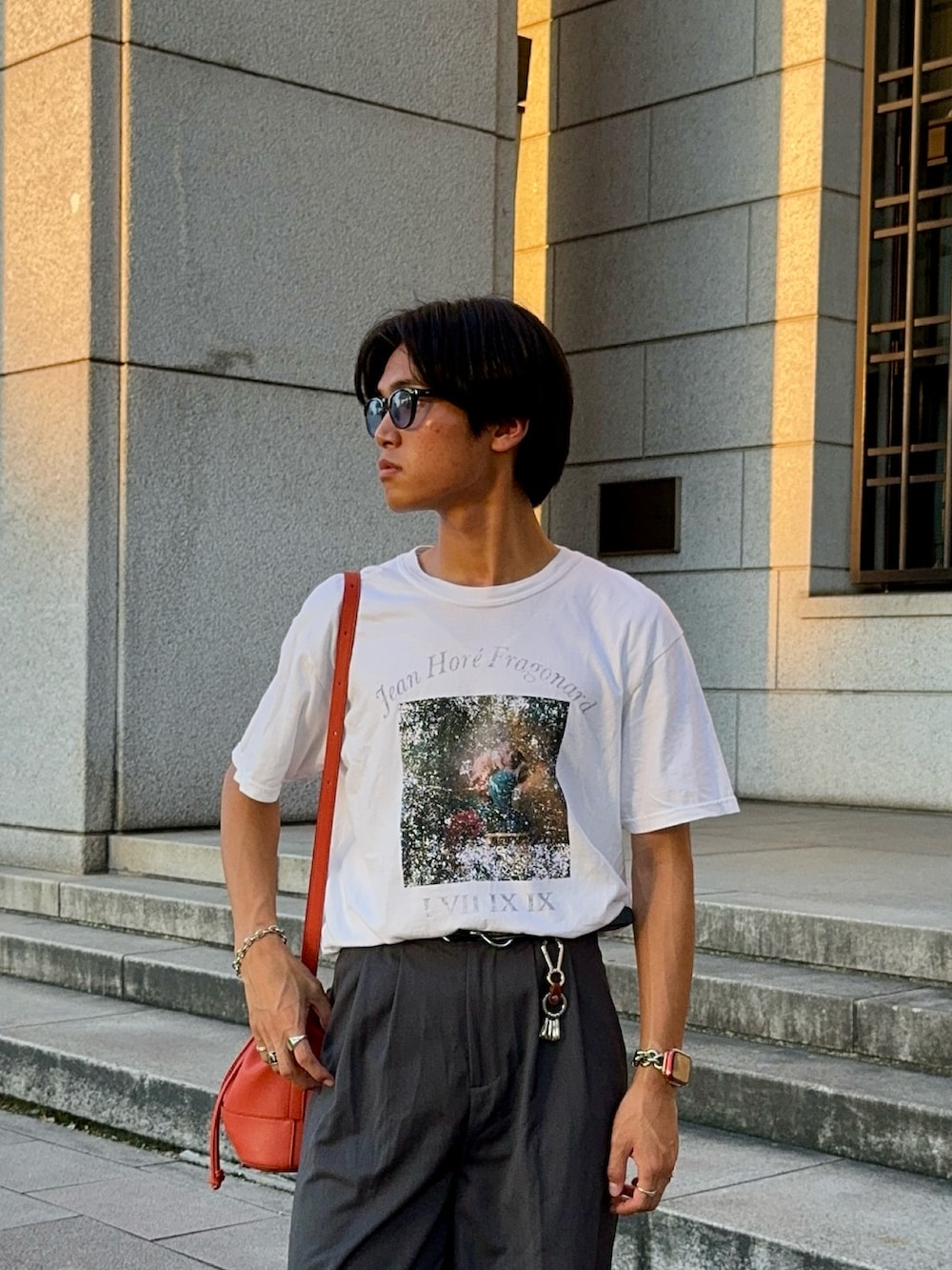 ホワイト系のTシャツ/カットソー、オレンジ系のバッグ、グレー系のスラックスを着用したメンズの夏コーディネートの2枚目の写真