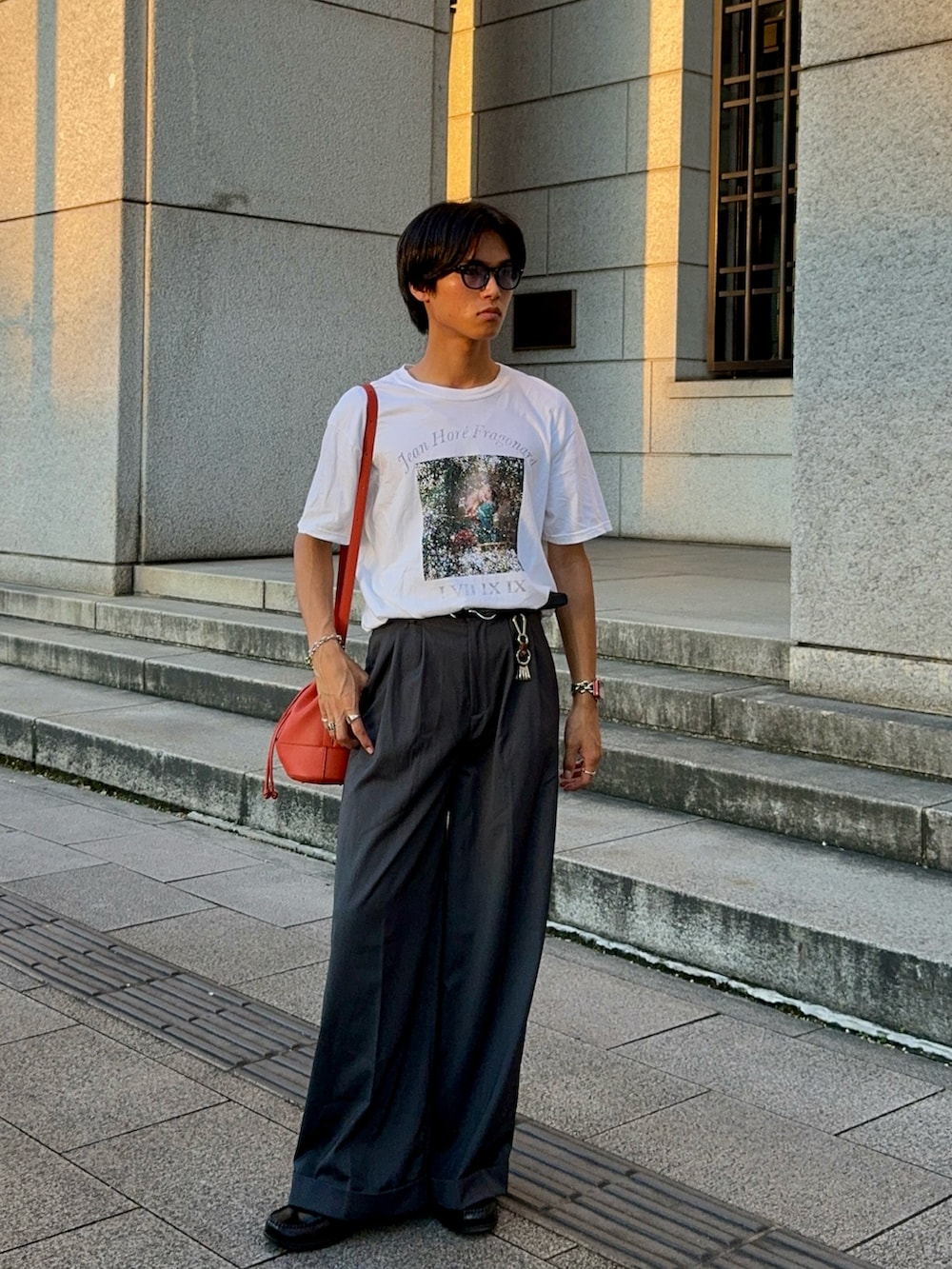 ホワイト系のTシャツ/カットソー、オレンジ系のバッグ、グレー系のスラックスを着用したメンズの夏コーディネートの5枚目の写真